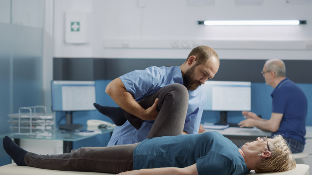 Un kiné pratique des étirements aux niveau de la jambe sur une patiente âgée. La femme est allongée sur une table de massage. 